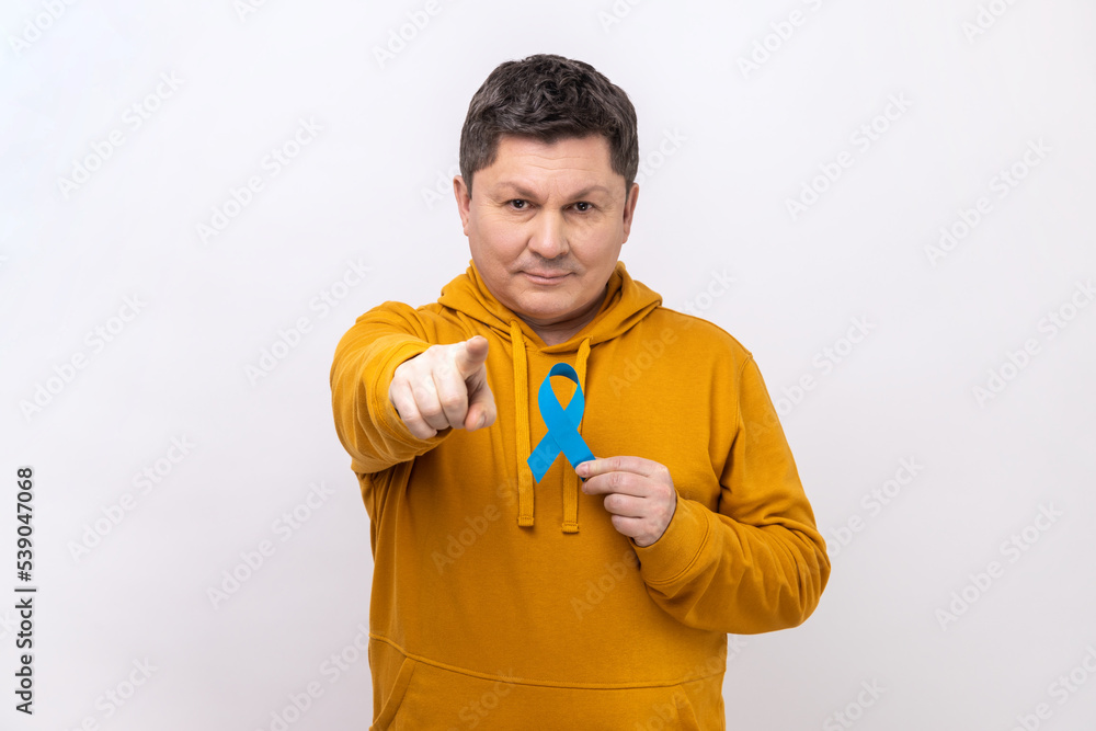 Portrait of serious attractive man holding blue awareness, disease symbol, looking and pointing at camera, support, wearing urban style hoodie. Indoor studio shot isolated on white background. Portrait of serious attractive man holding blue awareness, disease symbol, looking and pointing at camera, support, wearing urban style hoodie. Indoor studio shot isolated on white background.