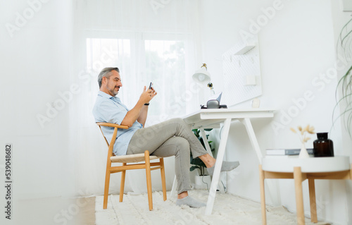 Mature adult man sitting at workplace holding mobile phone while working on laptop computer at home.