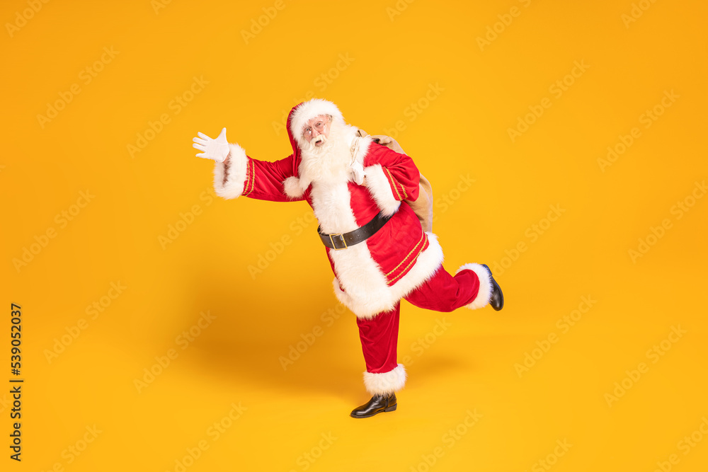 Merry Christmas and a happy new year ! Real Santa Claus carrying heavy sack with gifts, running fast on yellow studio background . Xmas time. Merry Christmas and a happy new year ! Real Santa Claus carrying heavy sack with gifts, running fast on yellow studio background . Xmas time.