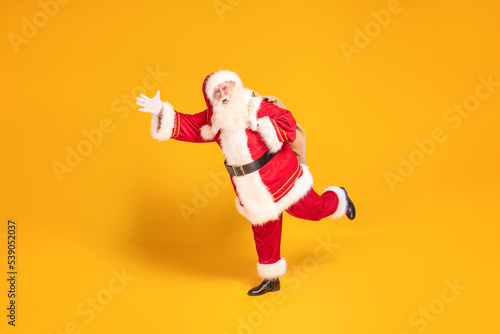 Merry Christmas and a happy new year ! Real Santa Claus carrying heavy sack with gifts, running fast on yellow studio background . Xmas time.