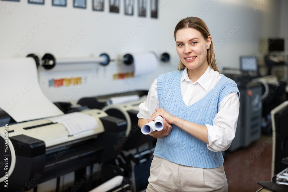 Portrait of positive woman printshop worker holding rolled pieces of paper and looking at camera.