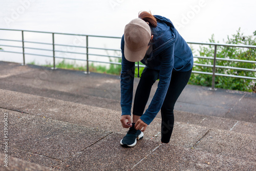 A woman wearing long-sleeved workout clothes bends down to tie her shoelaces in preparation for jogging.