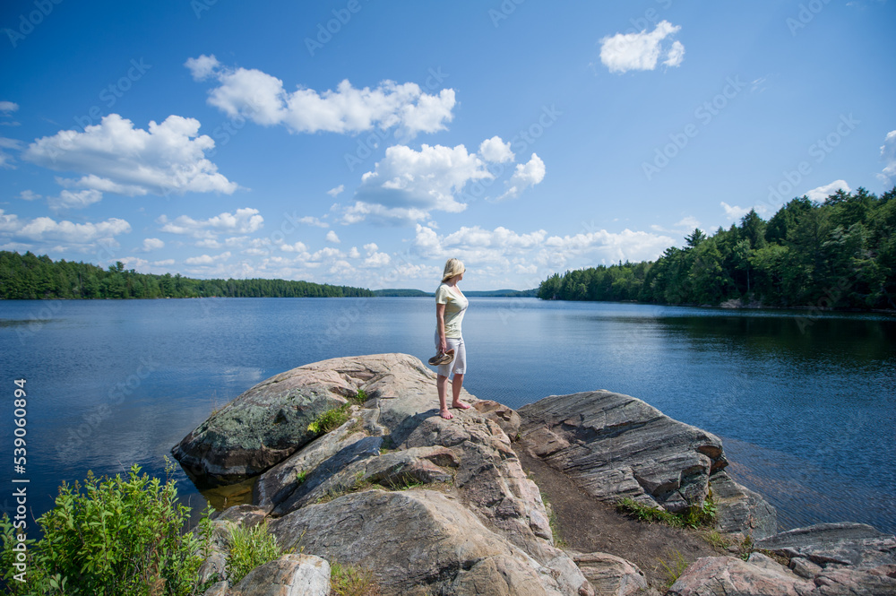 Young woman standing on a rock looking at Smoke lake in Algonquin Provincial Park, Muskoka Ontario Canada. The blue sky is showcasing beatiful fluffy clouds