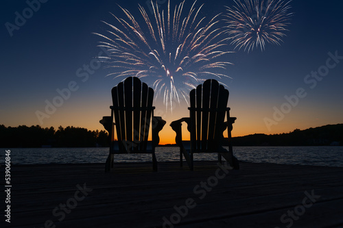Canada Day fireworks over two Adirondack chairs  on the wooden dock in Muskoka, Ontario Canada, are facing the sunset orange hues  while facing the calm water.