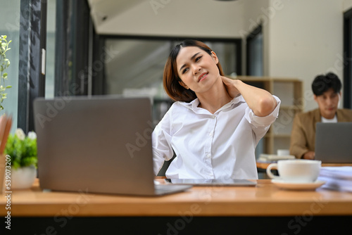 Exhausted and overworked Asian businesswoman suffering from neck pain at her desk.