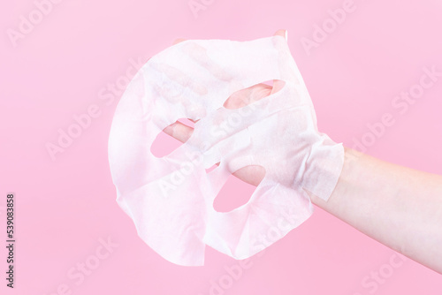 Female hand holding a white sheet of moisturizing mask close-up on pink background.