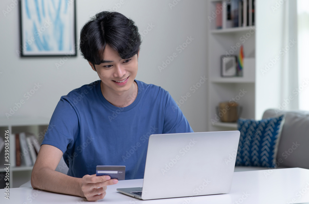 Young Asian man holding credit card and using laptop computer. Businessman working at home. Online shopping, e-commerce, internet banking, spending money, working from home concept Young Asian man holding credit card and using laptop computer. Businessman working at home. Online shopping, e-commerce, internet banking, spending money, working from home concept