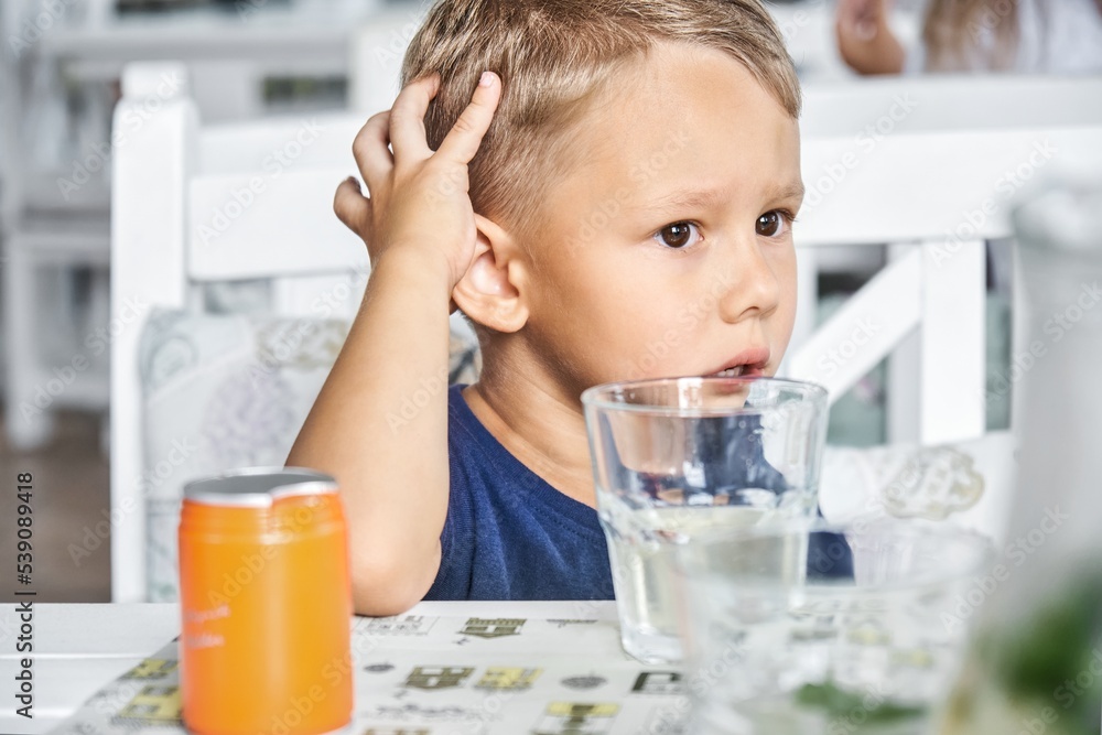 Preschooler boy sits in restaurant waiting for order to start eating. Fair-haired child looks with bored expression sitting at wooden table Preschooler boy sits in restaurant waiting for order to start eating. Fair-haired child looks with bored expression sitting at wooden table