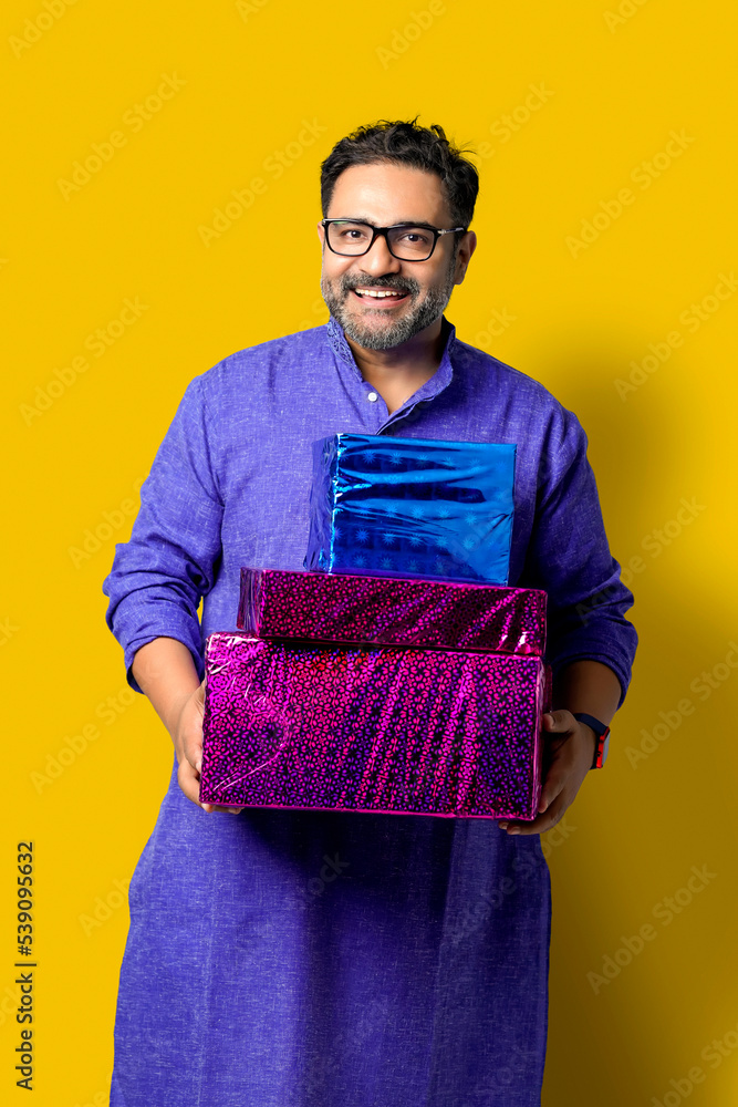 Young happy Indian man holding gift box and wearing traditional cloths, standing against yellow background Young happy Indian man holding gift box and wearing traditional cloths, standing against yellow background