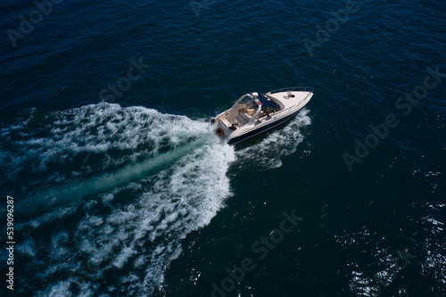 Big boat in motion on the water top view. Luxurious large yacht with people moving quickly on dark blue water making a white trail behind the boat.
