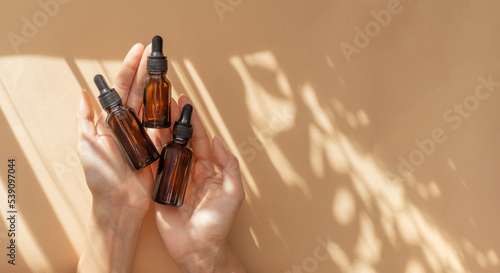 Woman's hand holds three amber glass vials with pipette on beige background in rays of sunlight. Top view of unmarked containers with essential oil, moisturizing serum. Cosmetic product for body care