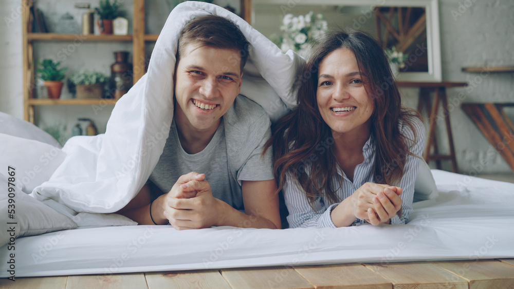 Portrait of happy couple lying in bed under blanket looking at camera laughing and smiling. Loving married people and happiness concept. Portrait of happy couple lying in bed under blanket looking at camera laughing and smiling. Loving married people and happiness concept.