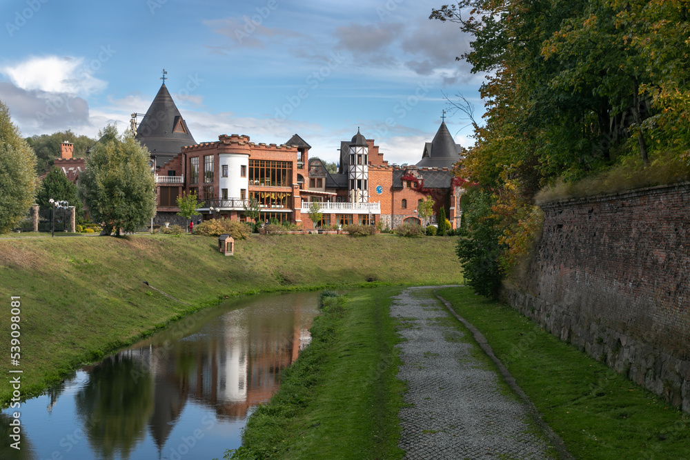 Cozy embankment of the city water canal. Ruins of a brick wall and a stone path along the river. Stream reflects an imitation of a medieval castle with pointed domes. Lots of trees and greenery around