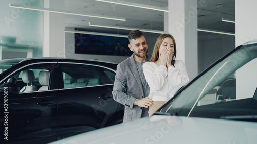 Attractive young woman is getting new car from her loving boyfriend  he is closing her eyes and leading her to auto in motor dealership  they are hugging and kissing.