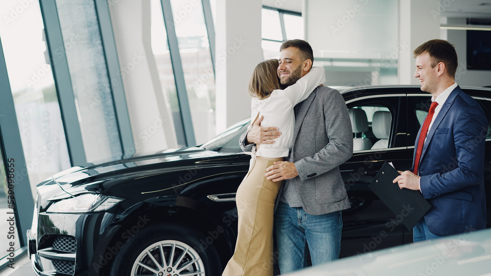 Young bearded man is buying car for his attractive wife, she is kissing and hugging him. Young bearded man is buying car for his attractive wife, she is kissing and hugging him.