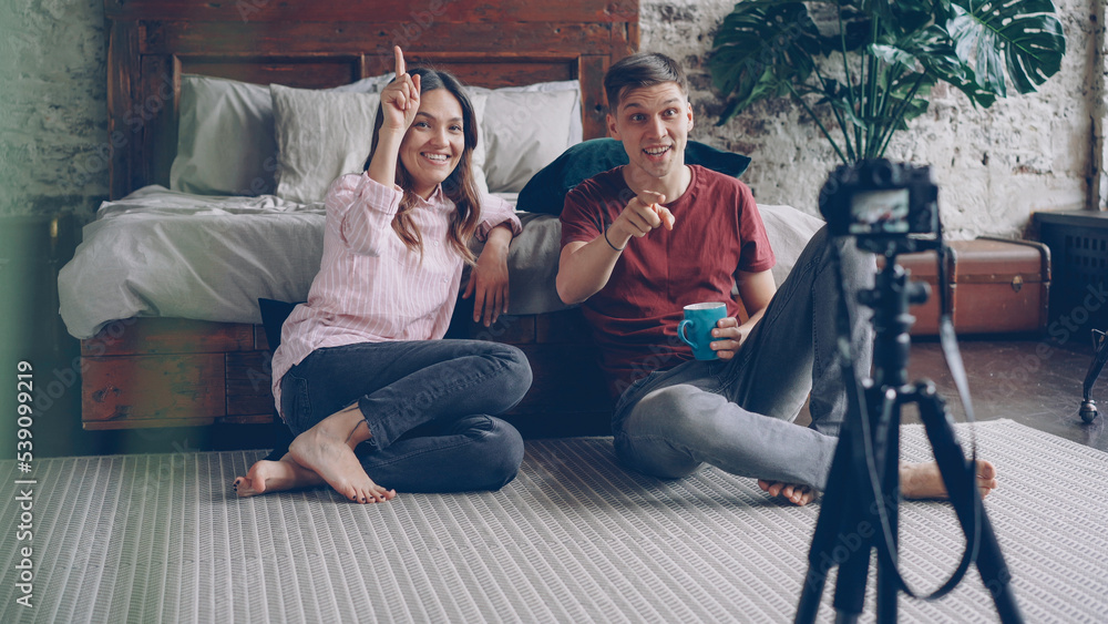 Naklejka premium Attractive young husband and wife bloggers are recording video vlog, waving hands and gesturing, talking and laughing, they are using camera on tripod and sitting on bedroom floor.