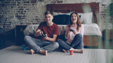 Cute young couple boyfriend and girlfriend are playing videogame holding joysticks sitting on bedroom floor at home. Emotional lovers are enjoying computer game.
