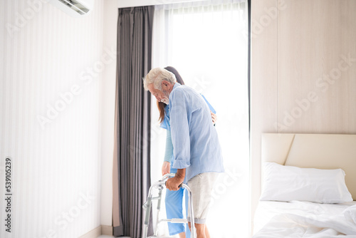 Portrait of Asian young nurse helping old elderly disable man grandfather to walk by using walker equipment in the bedroom. Senior patient of nursing home moving with walking frame and nurse support