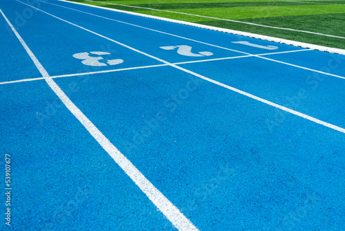 Blue running track in outdoor stadium with blue asphalt or rubber and white markings for fitness or competition with green grass field.