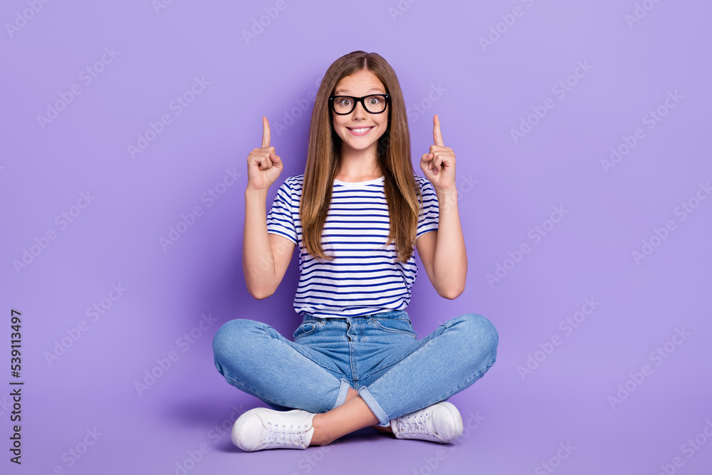 Full length photo of impressed girl striped t-shirt jeans sitting on floor hold directing empty space isolated on purple color background Full length photo of impressed girl striped t-shirt jeans sitting on floor hold directing empty space isolated on purple color background