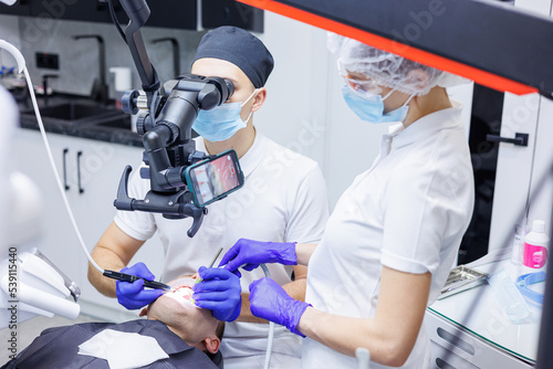 A young dentist looks at the patient's teeth with a dental microscope and holds dental instruments near the mouth. An assistant helps the doctor. They wear white uniforms with masks