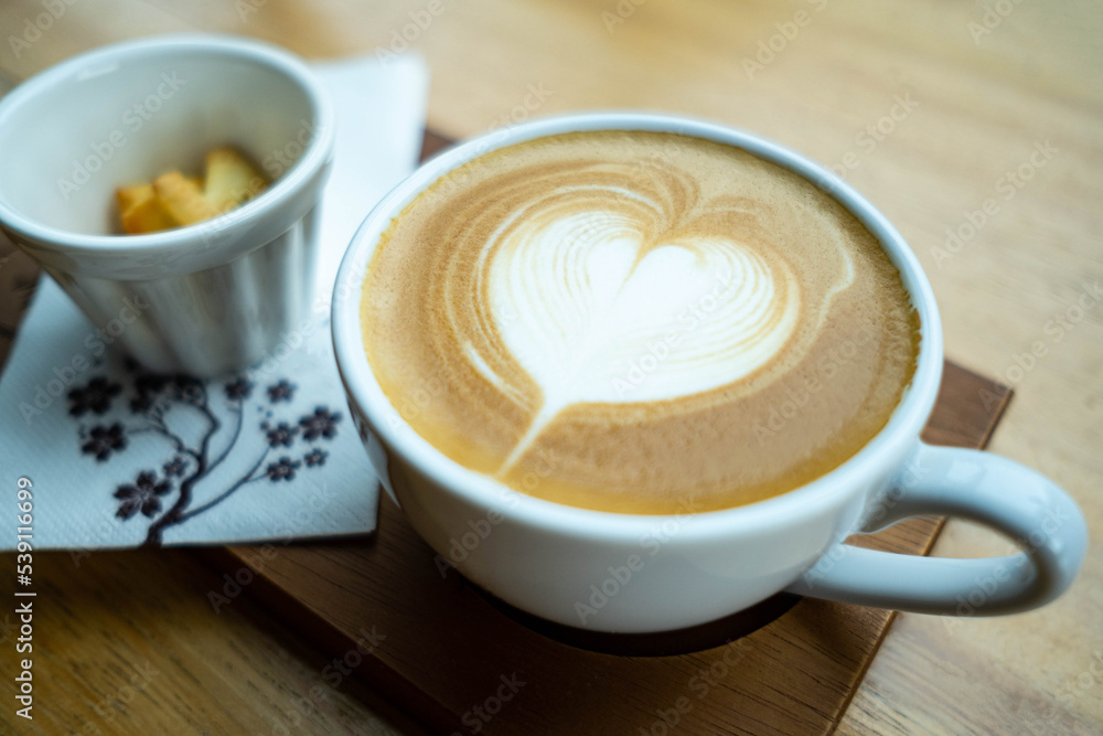 A white cup of latte art coffee with heart shape on old wooden background and there is a cup of biskit beside