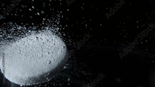 Macro shot of white tablet of soluble aspirin bubbling in glass of water on dark background. Effervescent tablet of acetylsalicylic acid dissolves in water forming many hiss bubbles. Headache pill. photo