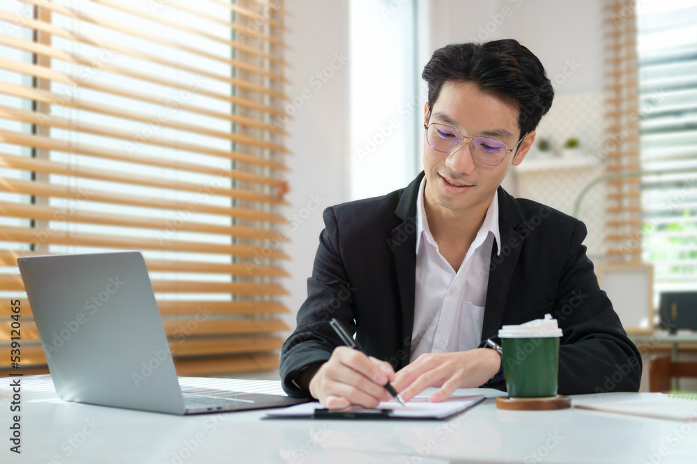 Focused asian businessman writing important information on notepad and using laptop at working desk. Focused asian businessman writing important information on notepad and using laptop at working desk.