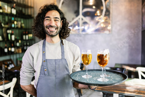 Friendly portrait of confident young adult waiter male holding serving tray with beers smiling at camera at brewery bar