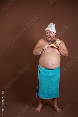 A fat man in a blue towel and a bath hat. Rest in the sauna. Happy guy on a brown background.