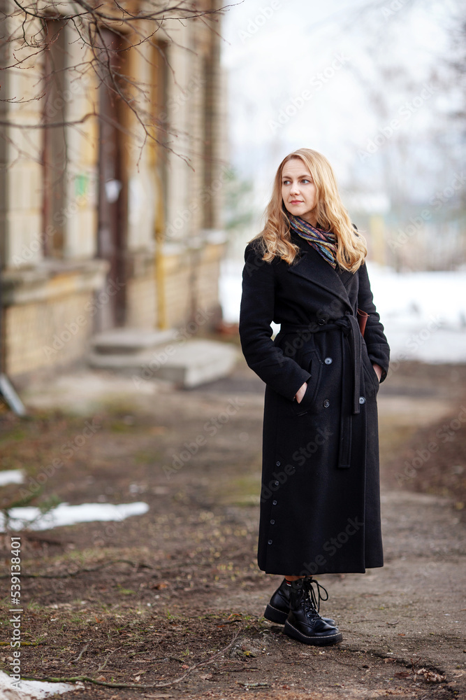Elegant young woman with long blonde hair in a black coat poses in the city. Urban portrait of a modern woman