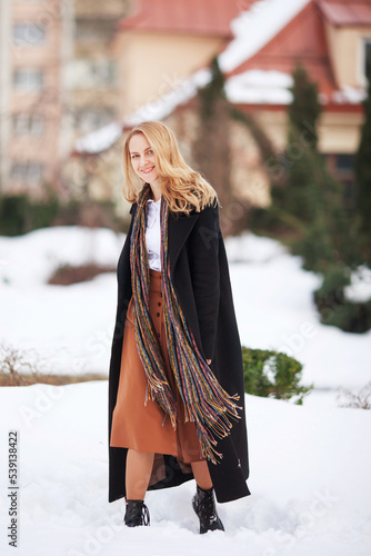 Elegant young woman with long blonde hair in a black coat poses in the city. Urban portrait of a modern woman