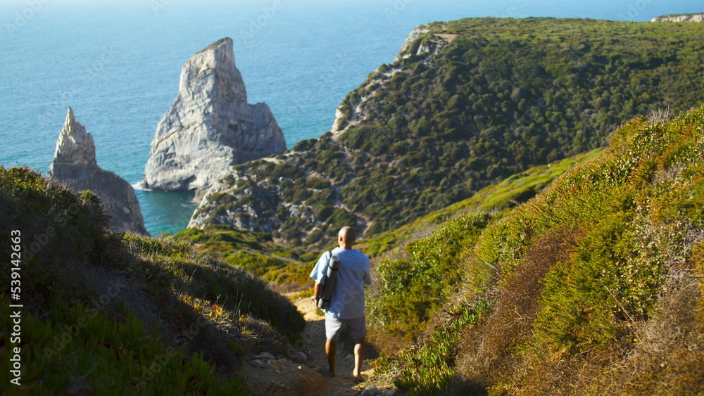 Man walking ocean cliff holding fitness mat on sunny day. Tourist going green
