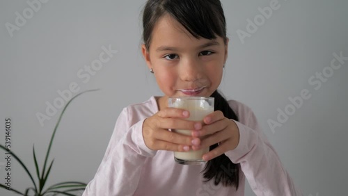 Little toddler girl drinking milk from the glass. Child drinks healthy yogurt and licks her lips. Positive kid having breakfast and enjoying natural fresh milk in the morning at home.