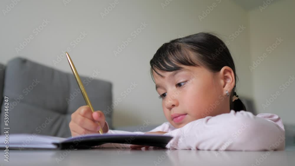 Little girl is writing the essay in notebook at home. Online learning, homeschooling. Kid is doing homework concept. Selective focus Little girl is writing the essay in notebook at home. Online learning, homeschooling. Kid is doing homework concept. Selective focus