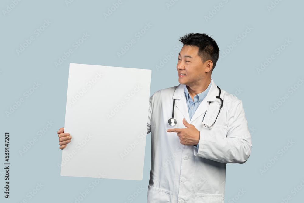 Smiling middle aged korean male in white coat with stethoscope points finger at banner with copy space Smiling middle aged korean male in white coat with stethoscope points finger at banner with copy space