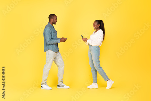 Cheerful African American Couple Using Mobile Phones Over Yellow Background