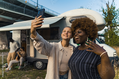 Two beautiful people in the foreground - queer caucasian person taking a selfie with black cellphone with their friendly Black friend in stripped dress. Traveling camper and male friends with guitar