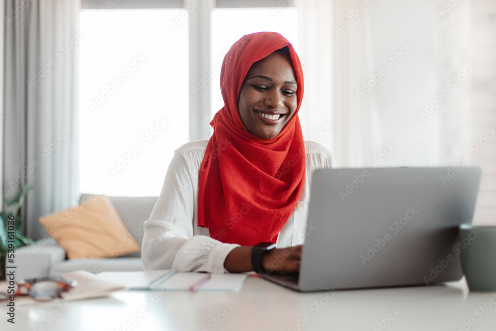 Remote job. Happy islamic woman in red hijab working on laptop, typing on keyboard and looking at computer screen