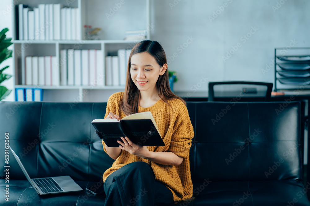 Fototapeta premium Portrait of a smiling Asian woman sitting on a sofa. Using laptop in the living room at home, working at home via online video conference.