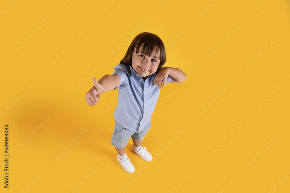 Above view portrait of little boy showing thumb up gesture, smiling to camera, orange studio background, full length