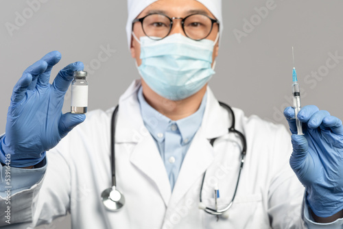 Calm mature chinese male doctor in white coat, protective gloves, mask and glasses show vaccine and syringe