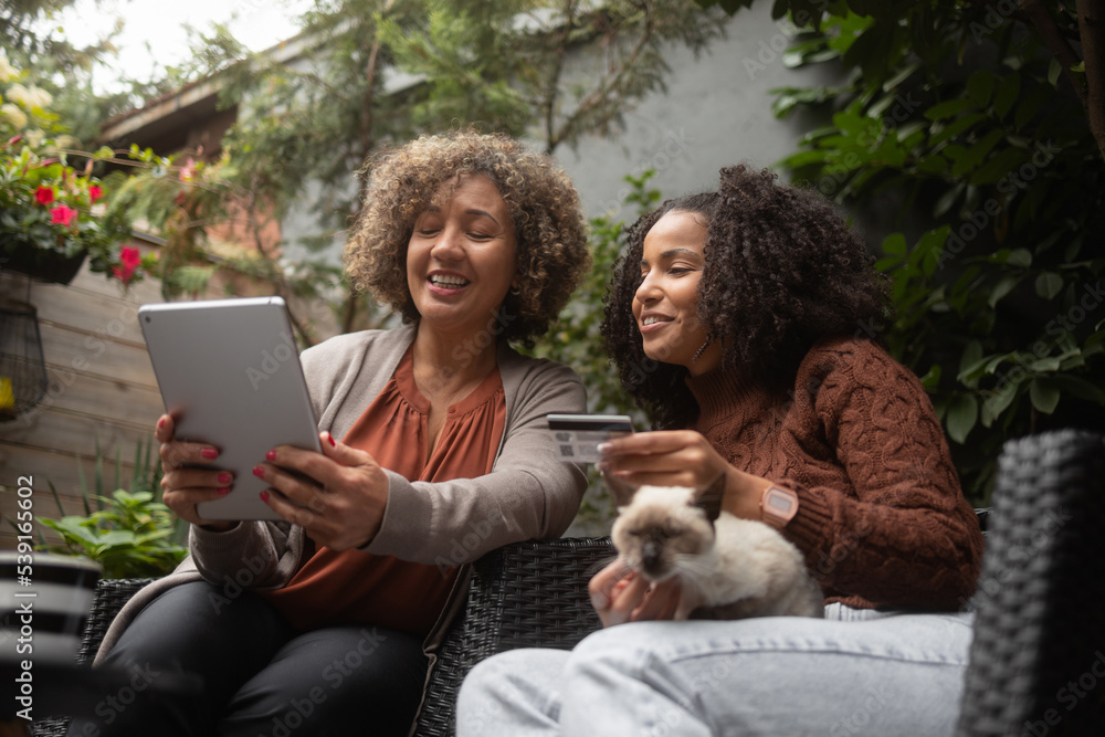 An African-American woman and her daughter use a digital tablet for online shopping. They sit on the patio and drink a coffee. The daughter is petting her siamese cat. An African-American woman and her daughter use a digital tablet for online shopping. They sit on the patio and drink a coffee. The daughter is petting her siamese cat.