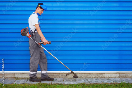 Young worker in overalls mows grass with trimer against background of blue metal siding. Janitor treats area from weeds. Workflow. Industrial background with space for copying..