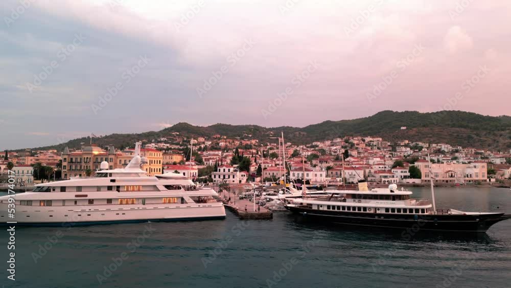 Flying backwards at the yacht pier in front of the Poseidonion Grand Hotel and main squar of Spetses revealing the cityscape and coast at sunset Flying backwards at the yacht pier in front of the Poseidonion Grand Hotel and main squar of Spetses revealing the cityscape and coast at sunset