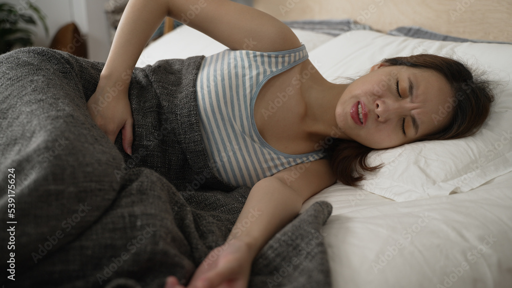 side view asian woman lying on white sheet of bed having stomach pain. sickness lady holding her painful belly while sleeping in comfort place at home. illness female holding abdomen ache in bedroom side view asian woman lying on white sheet of bed having stomach pain. sickness lady holding her painful belly while sleeping in comfort place at home. illness female holding abdomen ache in bedroom