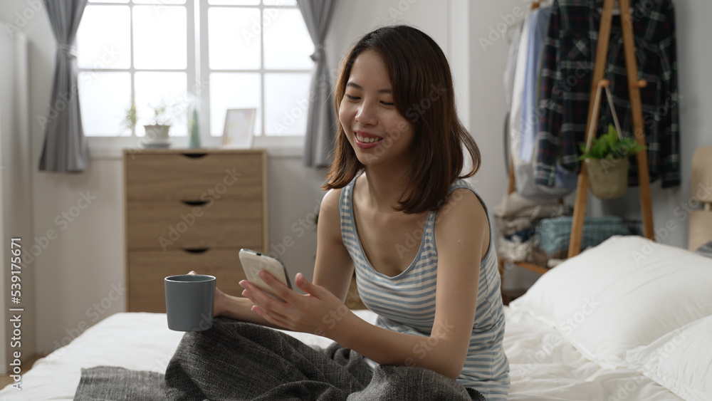attractive asian woman using smart phone at morning while drinking hot coffee in cozy bedroom. young lady sit in bed holding cup of tea and typing cellphone. girl enjoy breakfast and internet in bed attractive asian woman using smart phone at morning while drinking hot coffee in cozy bedroom. young lady sit in bed holding cup of tea and typing cellphone. girl enjoy breakfast and internet in bed