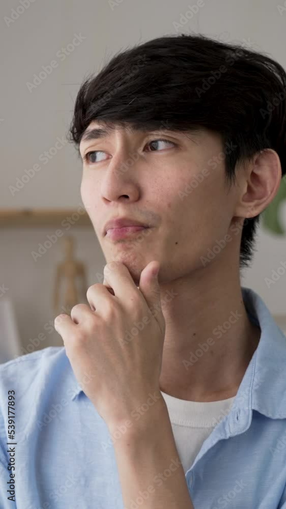 Vertical Screen: shoulder shot of a worried japanese guy looking into distance by the window while rubbing chin and thinking about a problem with anxiety at home Vertical Screen: shoulder shot of a worried japanese guy looking into distance by the window while rubbing chin and thinking about a problem with anxiety at home
