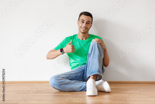Cheerful arab guy sitting on floor, smiling and showing thumb up, gesturing sign of approval against white wall