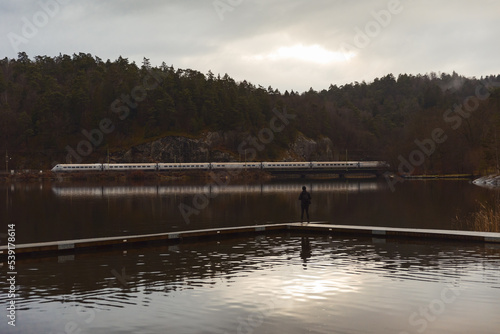 A male hiker standing on a bridge in a lake watching a train passing by.